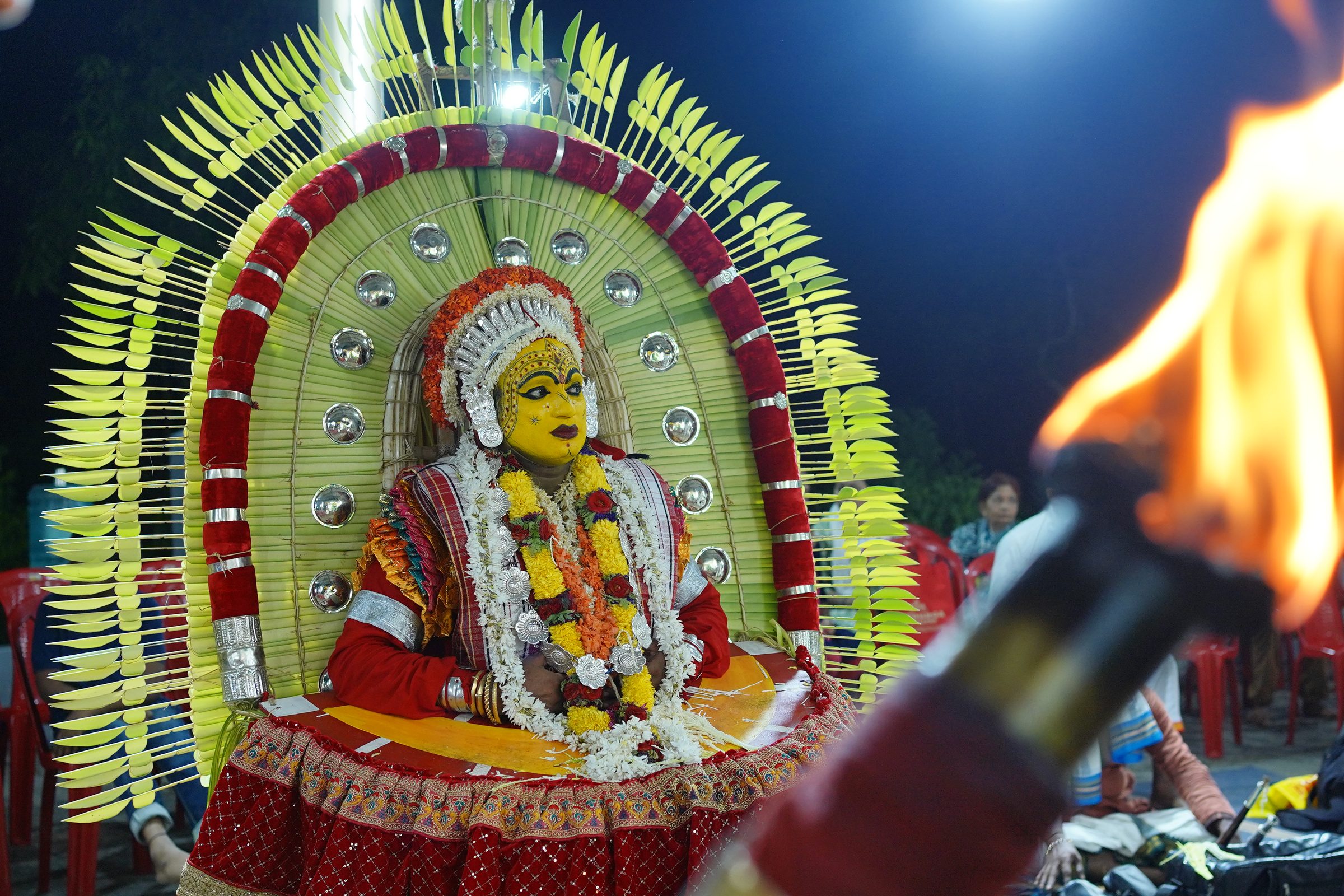A Kola performer in traditional attire beside the torch flame, at a night ritual of coastal Karnataka